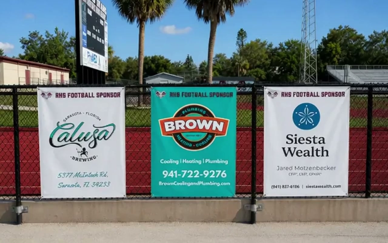Outdoor vinyl banners with grommets mounted on a chain-link fence at a sports field